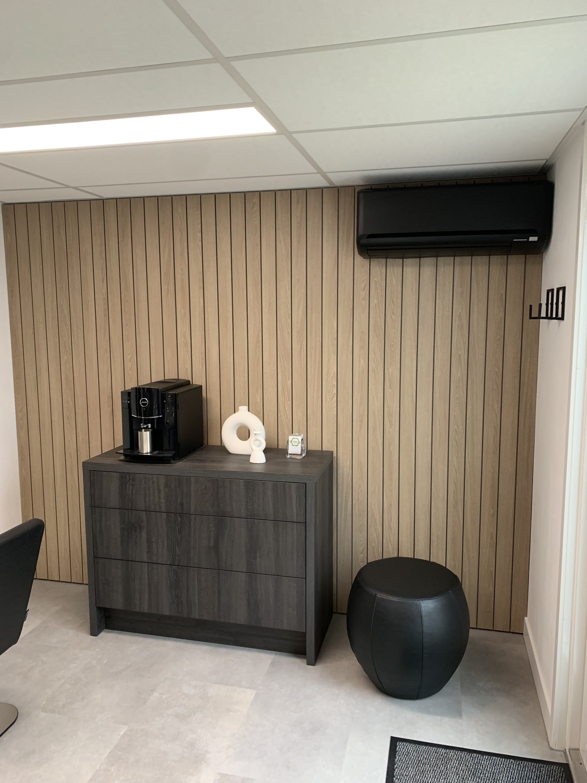 Hoek van de salon met houten lattenwand, een koffiemachine op een donkere kast en een zwarte kruk